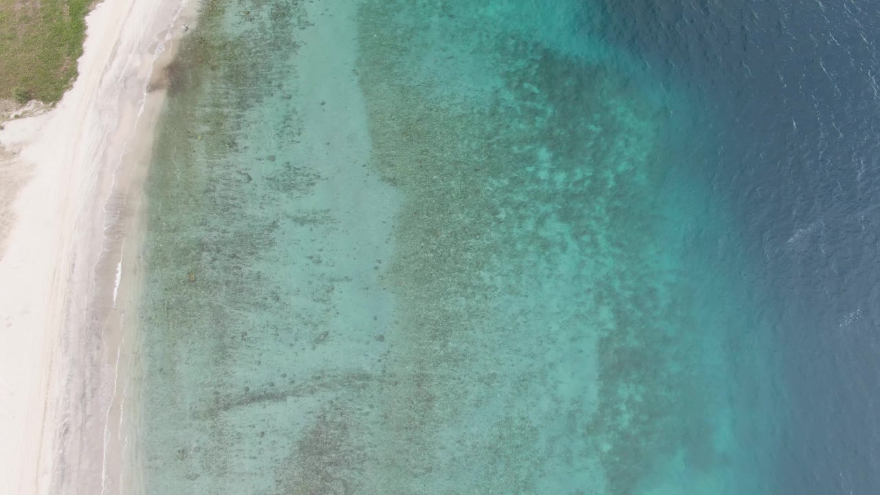 Aerial view over Los Barriles shoreline with the sandy beach slightly more defined at left, transparent turquoise water over mottled seabed, dark blue ocean at right, and no visible people.
