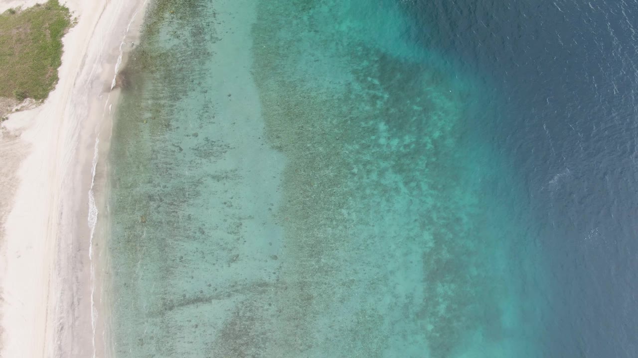 Aerial frame over the Los Barriles coast with pale sand running vertically along the left edge, clear green-blue shallows in the middle, darker offshore water to the right, and no visible people.