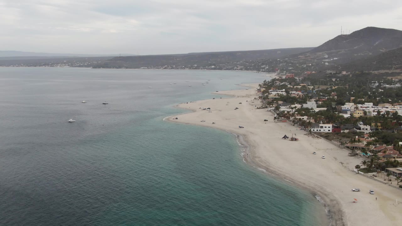 Drone-style aerial frame over Los Barriles, Baja California Sur, showing turquoise water beside a pale sand shoreline, a coastal road with vehicles, scattered buildings, distant hills, overcast sky, and no visible people.