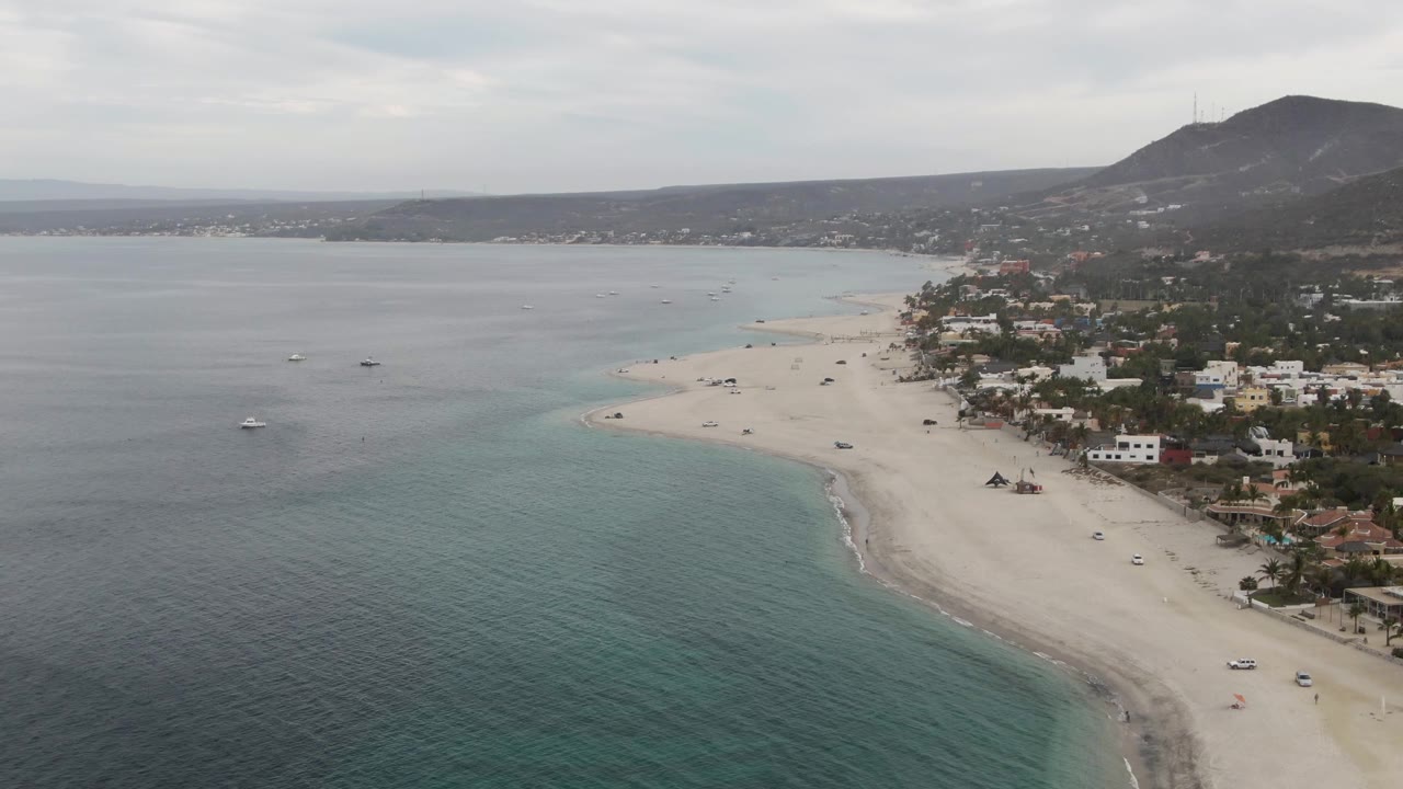 Wide aerial view of Los Barriles with turquoise shallows in the foreground, a curving sandy beach, roadside vehicles near scattered buildings, small boats offshore, background hills, gray clouds, and no visible people.