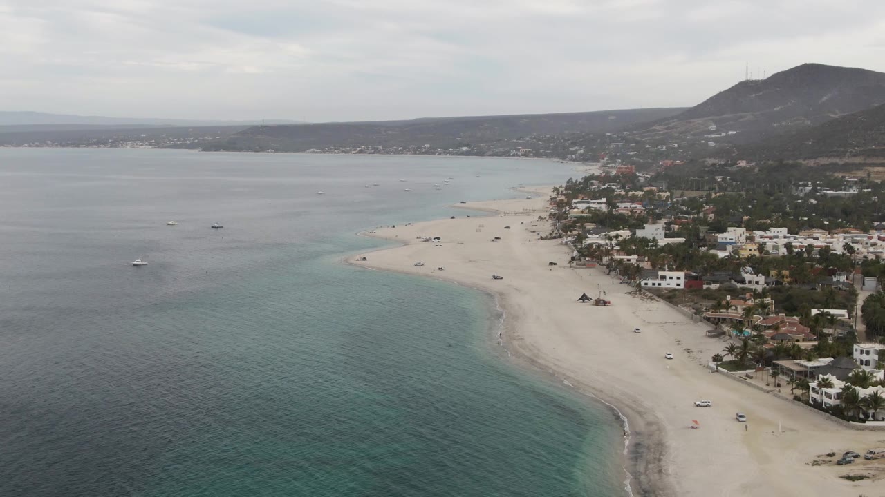 Aerial frame from Los Barriles, Baja California Sur showing turquoise water in the foreground, a pale sandy beach curving along shoreline homes, boats offshore, distant hills, and no visible people.