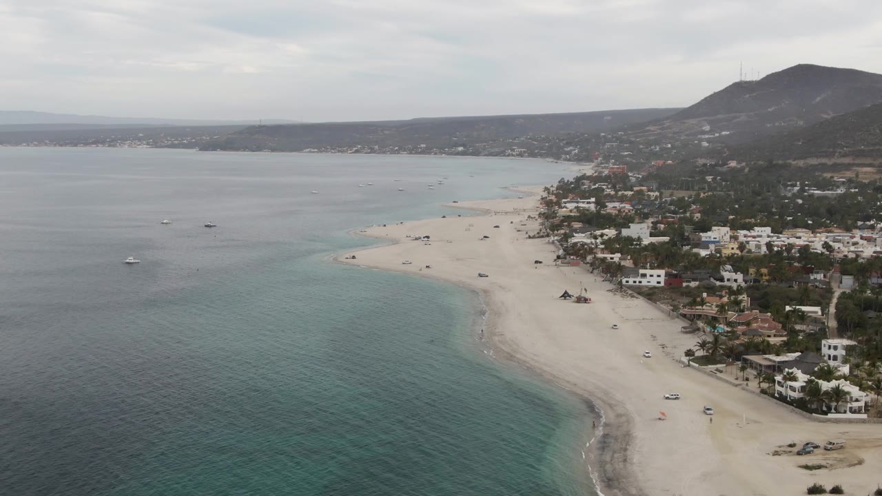 A Los Barriles aerial still with calm gray-blue water to the left, lighter turquoise shallows near the sand, beach vehicles, shoreline buildings, palms, a dark inland hill, and no visible people.