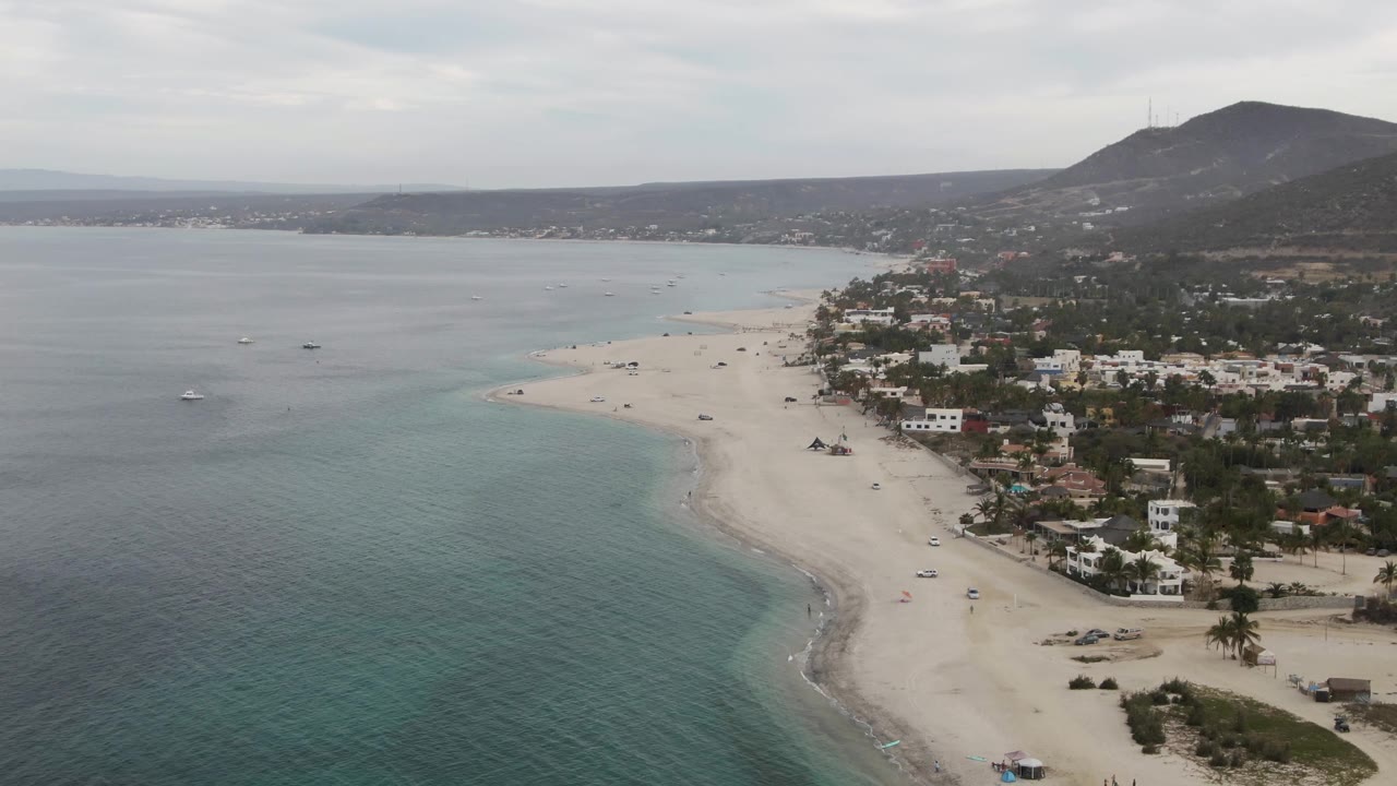 Aerial frame from Los Barriles showing a pale sandy shoreline, turquoise water in the foreground, low coastal buildings, palms, mountains, and tiny beachgoers near the shore.