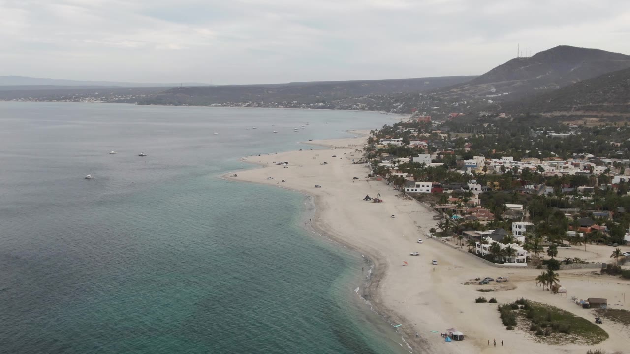 Drone view over clear water toward a sandy shoreline, beachgoers, buildings, and mountains.