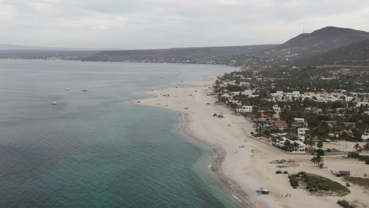 A high oblique frame over Los Barriles shows turquoise water at left and bottom, a pale beach sweeping along the town, tiny people on the sand, low buildings, palm trees, and a dark mountain ridge.