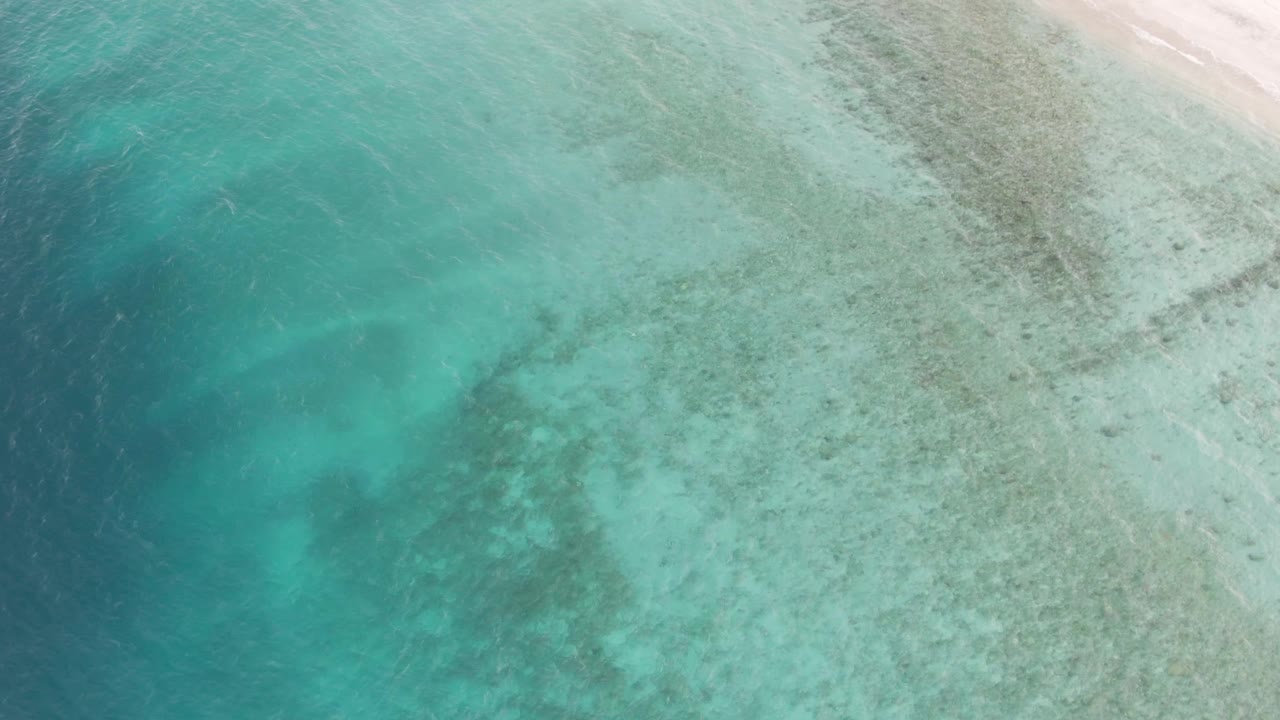 Aerial coastal view in Los Barriles with turquoise water, darker submerged patches, light ripples, and pale sand along the upper right edge, no visible people.