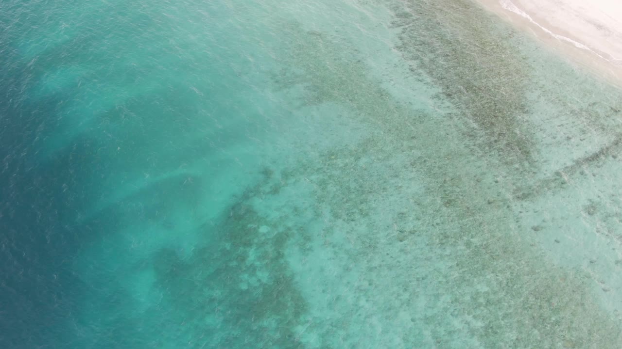 Mid-clip aerial frame over Los Barriles shallow water, with a broad teal area on the left, speckled seabed on the right, and pale sand at the upper right, no visible people.