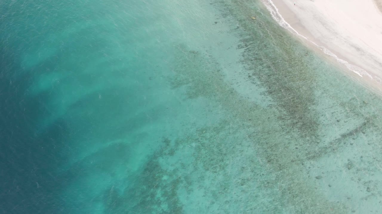 High aerial frame of turquoise coastal water, mottled seabed, and a pale beach strip at Los Barriles, Mexico, with no visible people.