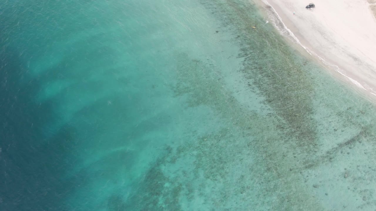 Drone-style view of clear turquoise water beside white sand in Los Barriles, with a small dark vehicle visible near the upper-right beach and no visible people.