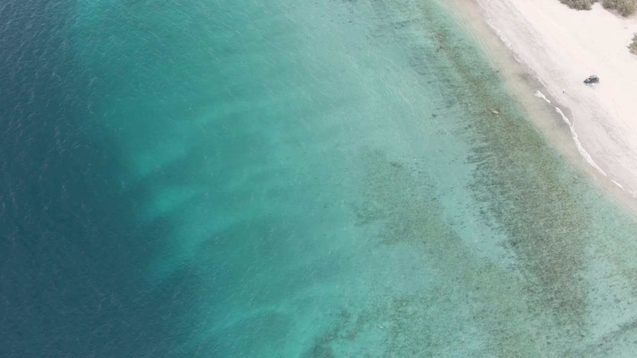 Aerial frame over Los Barriles coastline with a diagonal strip of pale beach at upper right, turquoise shallows across the center, darker water at left, and no visible people.
