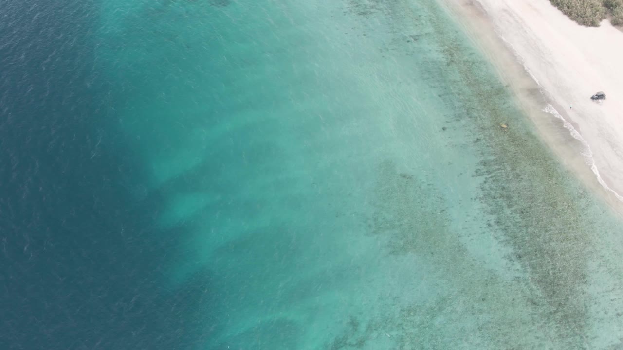 High overhead view in Los Barriles showing broad turquoise shallows, darker underwater patches near the beach, a small boat close to the sand at upper right, and no visible people.
