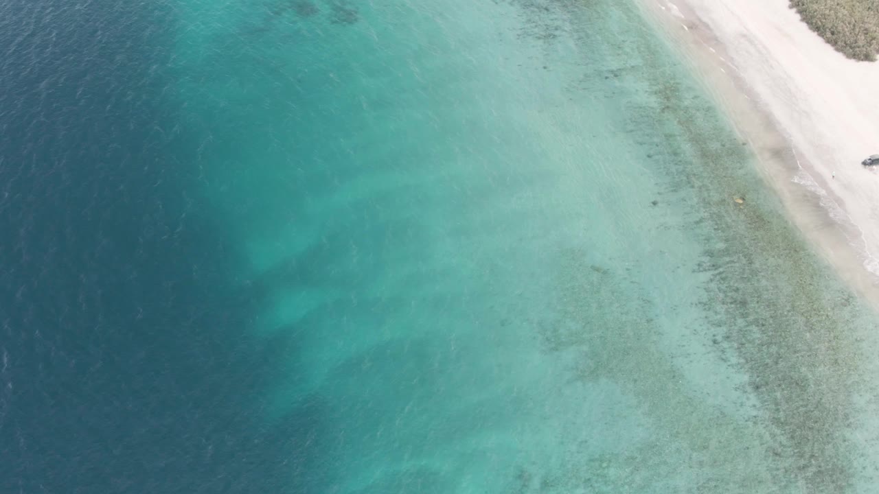 Mid-clip overhead frame from Los Barriles with the shoreline shifted lower in view, deep blue ocean on the left, clear shallow water across the center, pale sand at right, and no visible people.