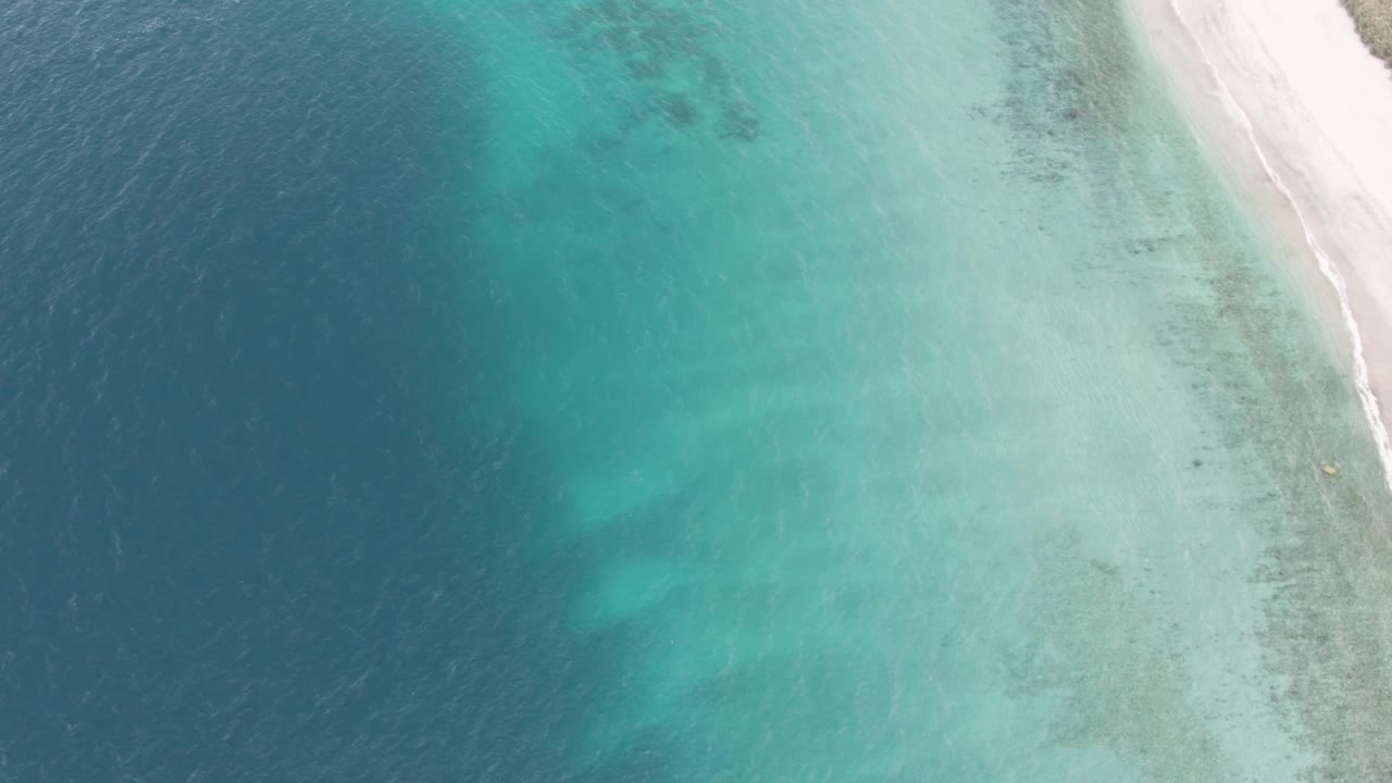 Aerial frame over Los Barriles clear coastal water, showing pale seabed through turquoise shallows and white sand along the right edge, with no visible people.