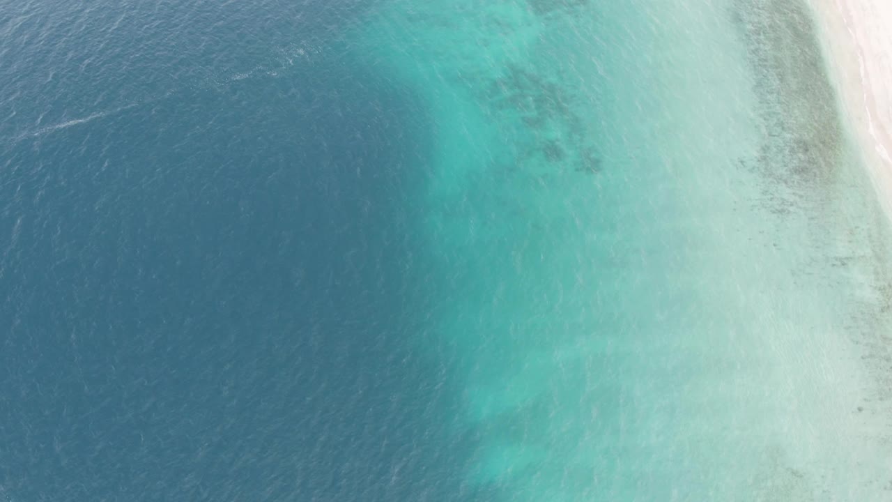 Overhead frame from Los Barriles shows dark blue ocean blending into turquoise shallows beside pale sandy shore, with no visible people.