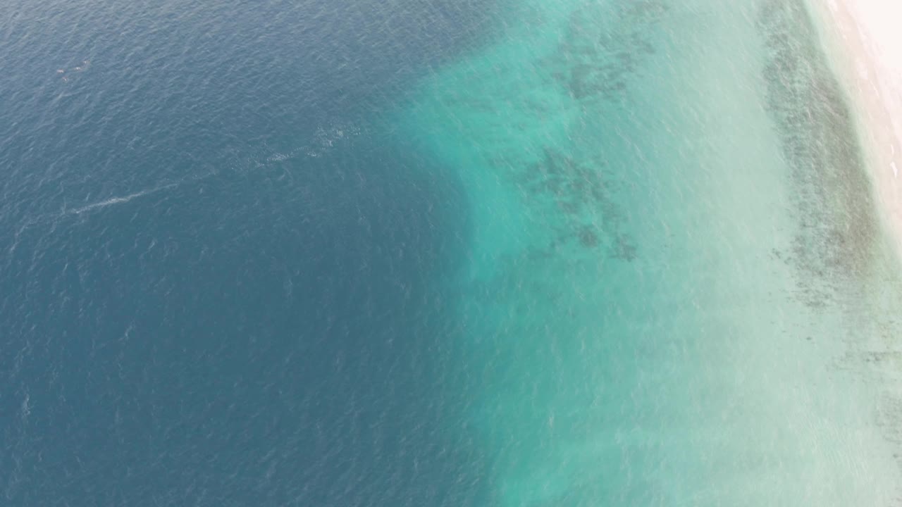 Drone-style overhead frame from Los Barriles shows shallow turquoise sea, darker reef-like patches under the surface, and a narrow pale beach, with no visible people.