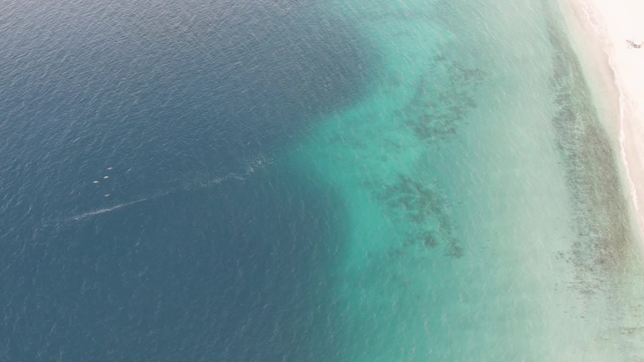 High aerial frame over Los Barriles coastal water, with deep blue ocean on the left, turquoise shallows near the sand edge, and a faint boat wake crossing the darker water.