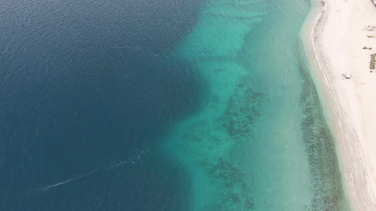 Aerial view of turquoise shallows, deep blue ocean, sandy beach, and a small vehicle near the shoreline.