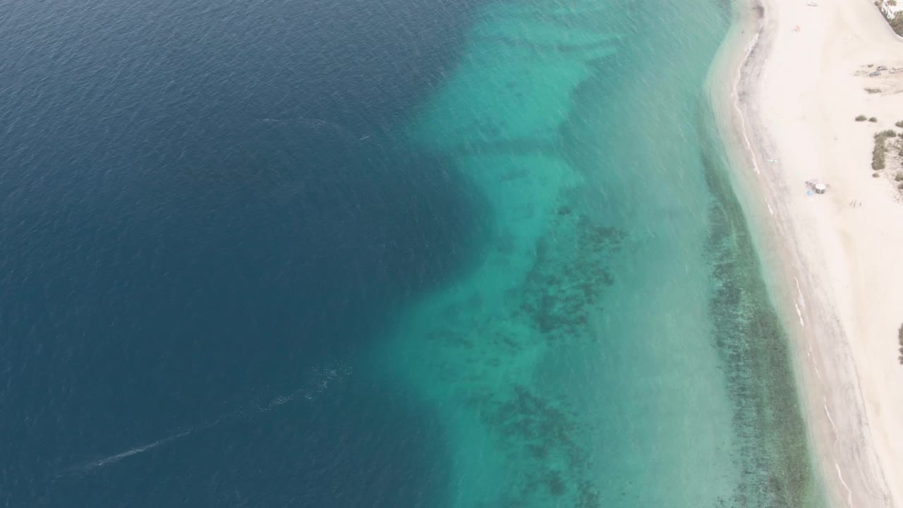 At 0:01, the Los Barriles aerial view keeps deep water on the left, clear shallows through the middle, pale beach on the right, the vehicle slightly farther along the shore, and no visible people.