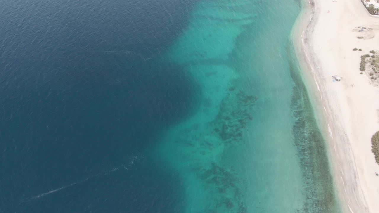 At 0:02, the overhead Los Barriles frame shows the shoreline curving through the right side, a small vehicle on the sand, transparent turquoise water over darker bottom patches, deep blue water offshore, and no visible people.
