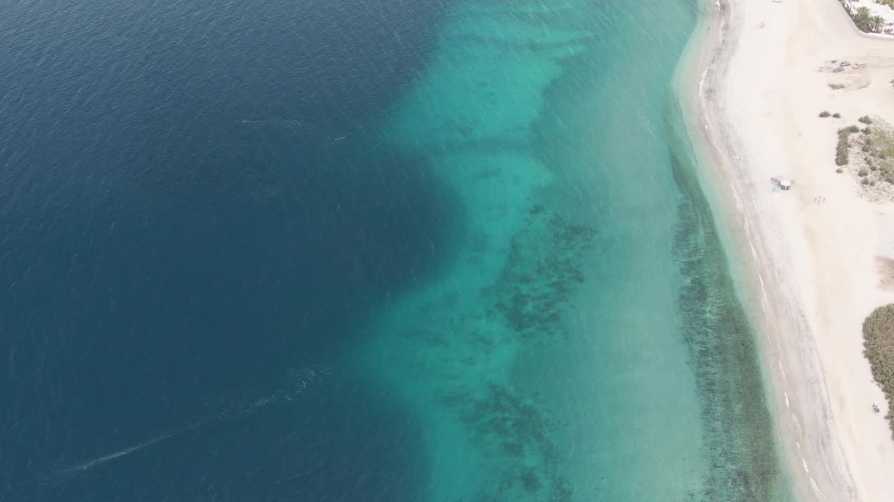 At 0:03, the Los Barriles shoreline remains viewed from above, with the vehicle continuing along the pale sand, darker seabed texture visible below the turquoise shallows, deep water at left, and no visible people.