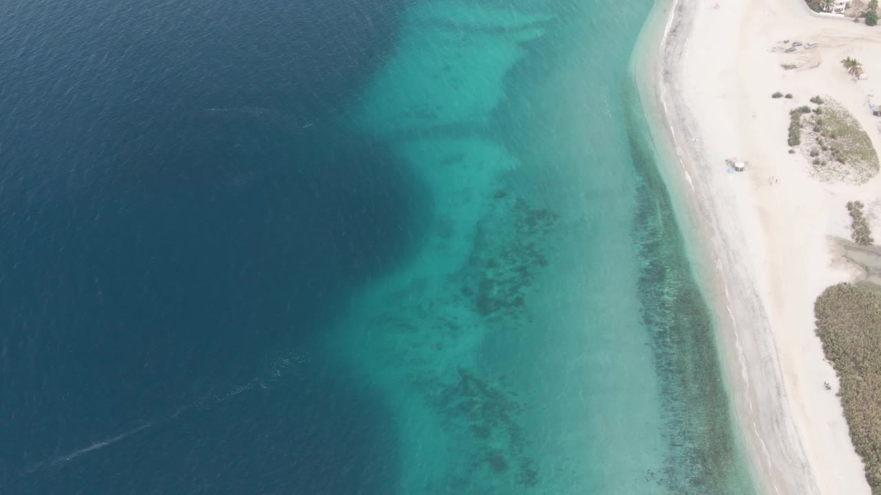 High overhead view in Los Barriles, Mexico, with deep blue ocean at left, turquoise shallows near shore, a pale sandy beach road at right, a small vehicle near the upper beach, and no visible people.
