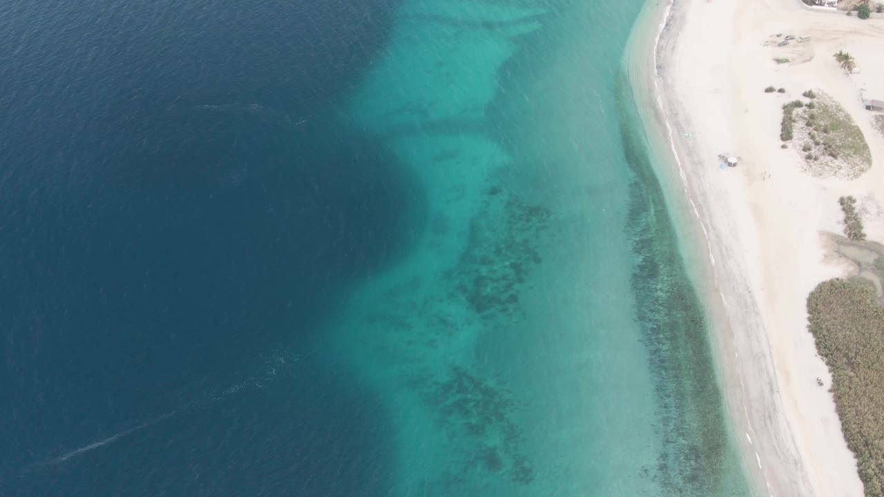 Aerial frame in Los Barriles, Mexico, showing the vehicle slightly farther along the sandy road, with dark ocean water on the left, clear turquoise shallows in the center, pale beach on the right, and no visible people.