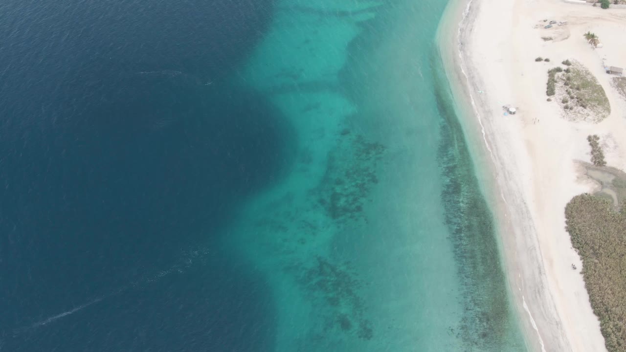 Los Barriles aerial view with the sandy shoreline pressed along the right edge, the vehicle near the upper beach road area, turquoise shallows below it, darker water offshore, and no visible people.