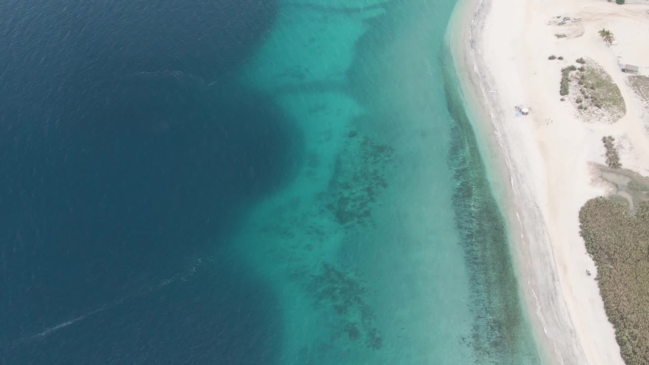 Aerial beach scene with deep blue water, turquoise shallows, sand, and a small vehicle.