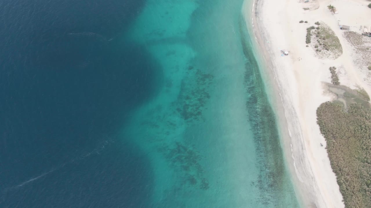 Drone view over Los Barriles showing a tiny vehicle near the upper right beach area, clear turquoise water, white sand, and no visible people.