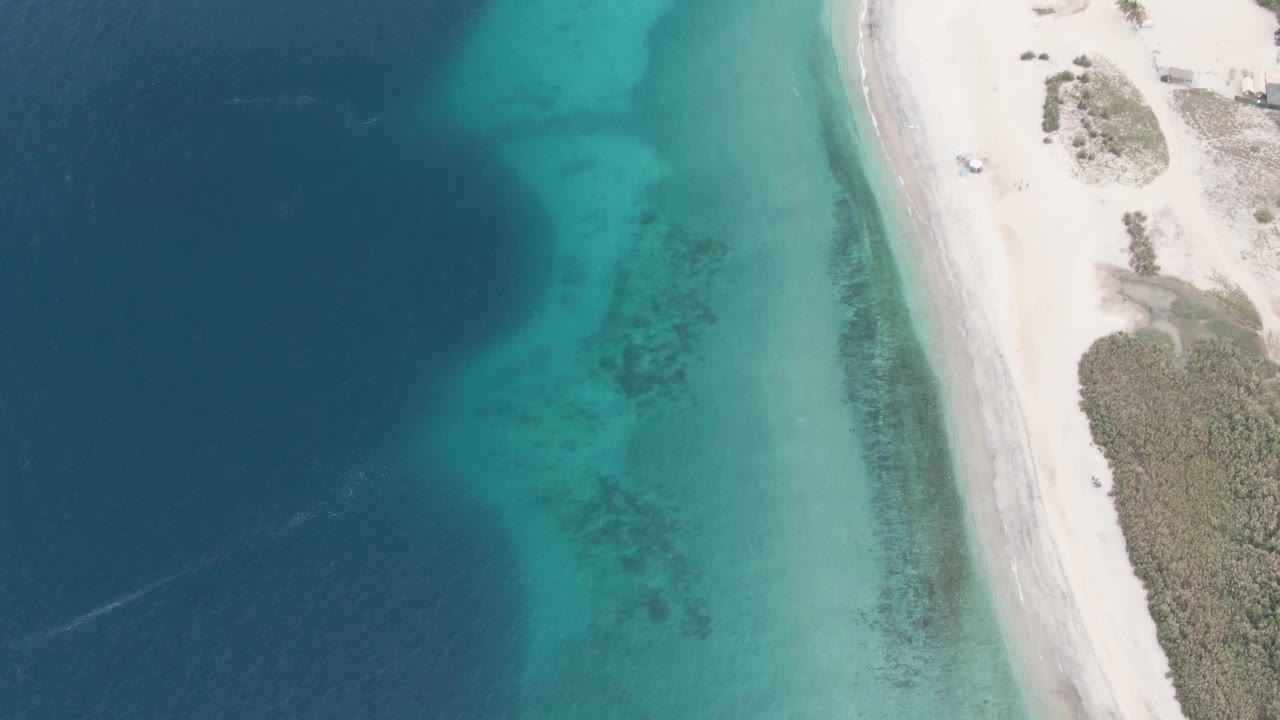 Aerial frame from Los Barriles with darker ocean on the left, turquoise shallows near shore, a narrow white beach on the right, and no visible people.
