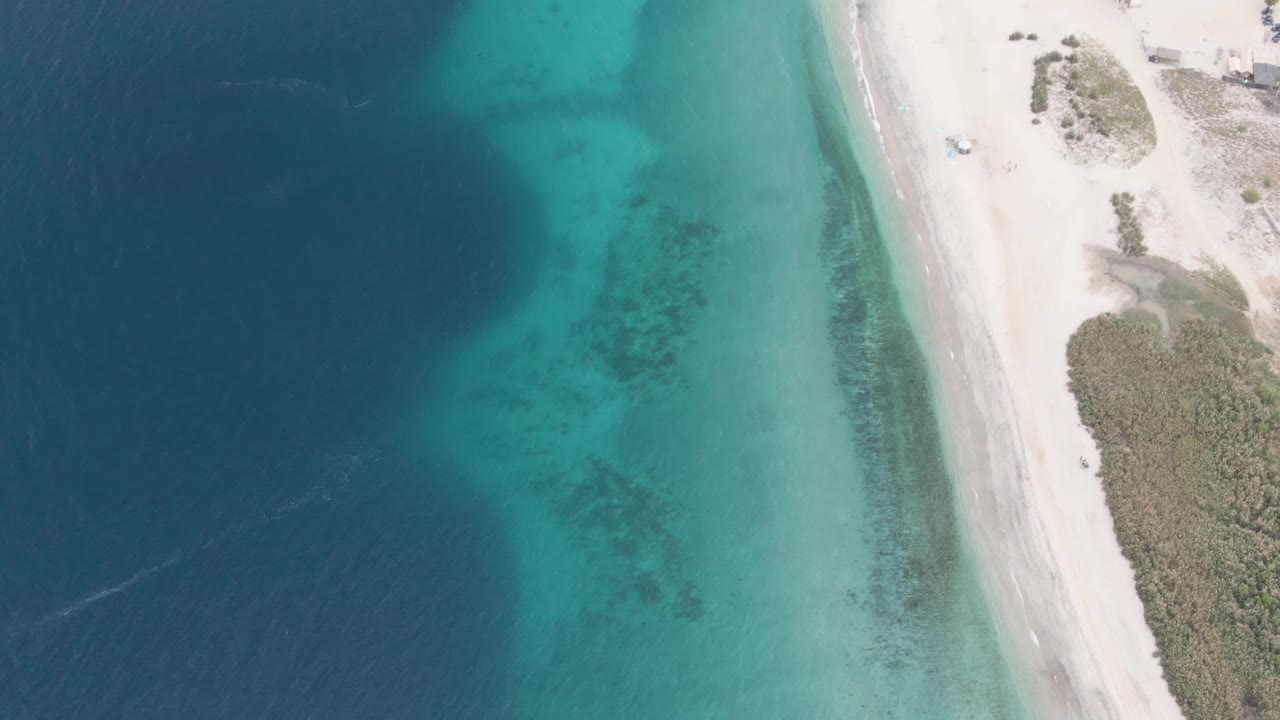 Mid-clip overhead view in Los Barriles where turquoise shallows broaden beside the pale sand beach, with scrub vegetation on the right and no visible people.