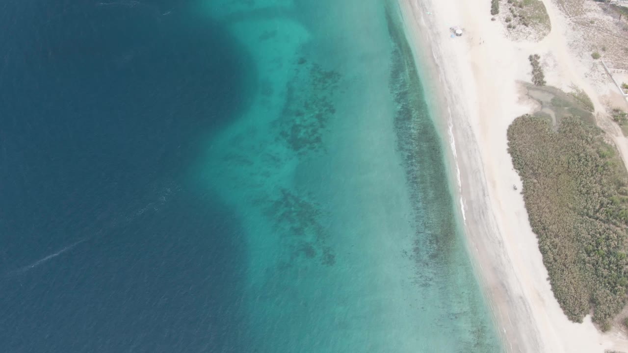 Aerial frame in Los Barriles, Baja California Sur, with the beach road running along the right edge, turquoise water in the center, darker offshore water at left, and no visible people.