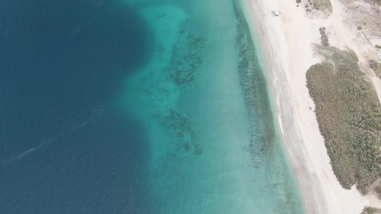 Overhead Los Barriles shoreline frame showing darker water at left, clear green-blue shallows near shore, pale beach sand, brushy vegetation to the right, and no visible people.