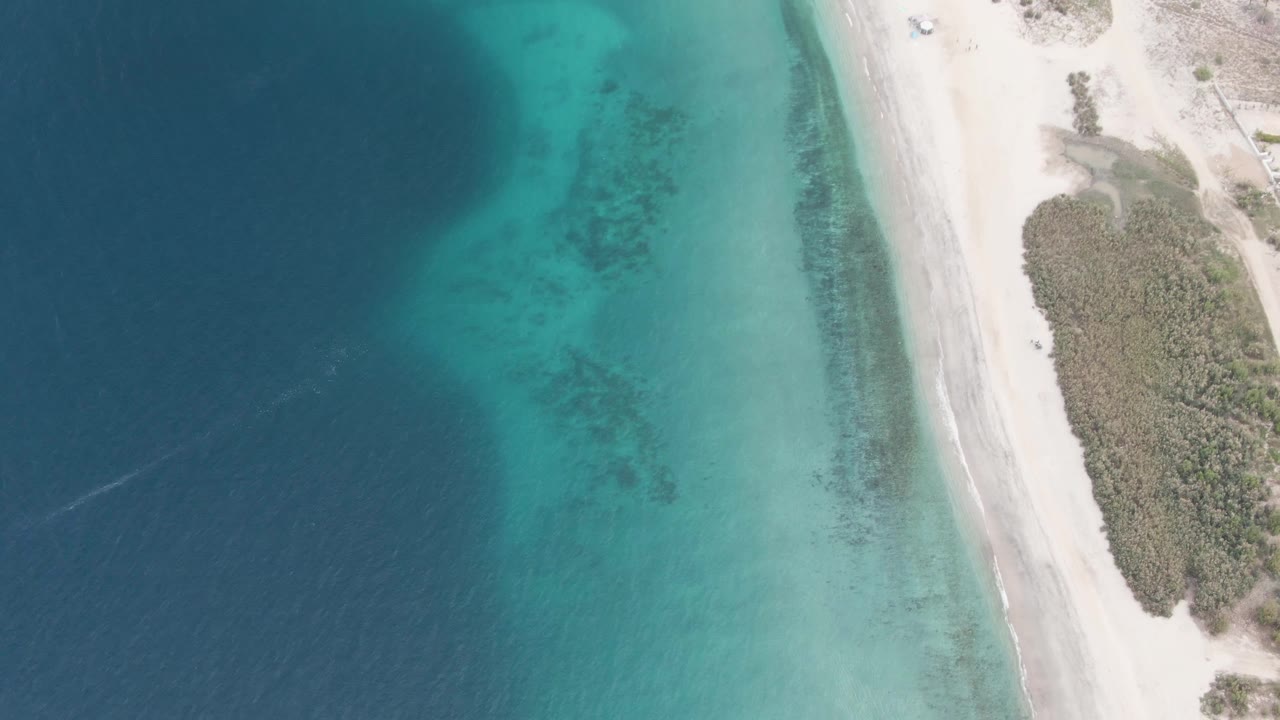 Mid-clip overhead view in Los Barriles, Baja California Sur, with a small vehicle near the upper sandy road, broad turquoise shallows below it, darker water to the left, and no visible people.
