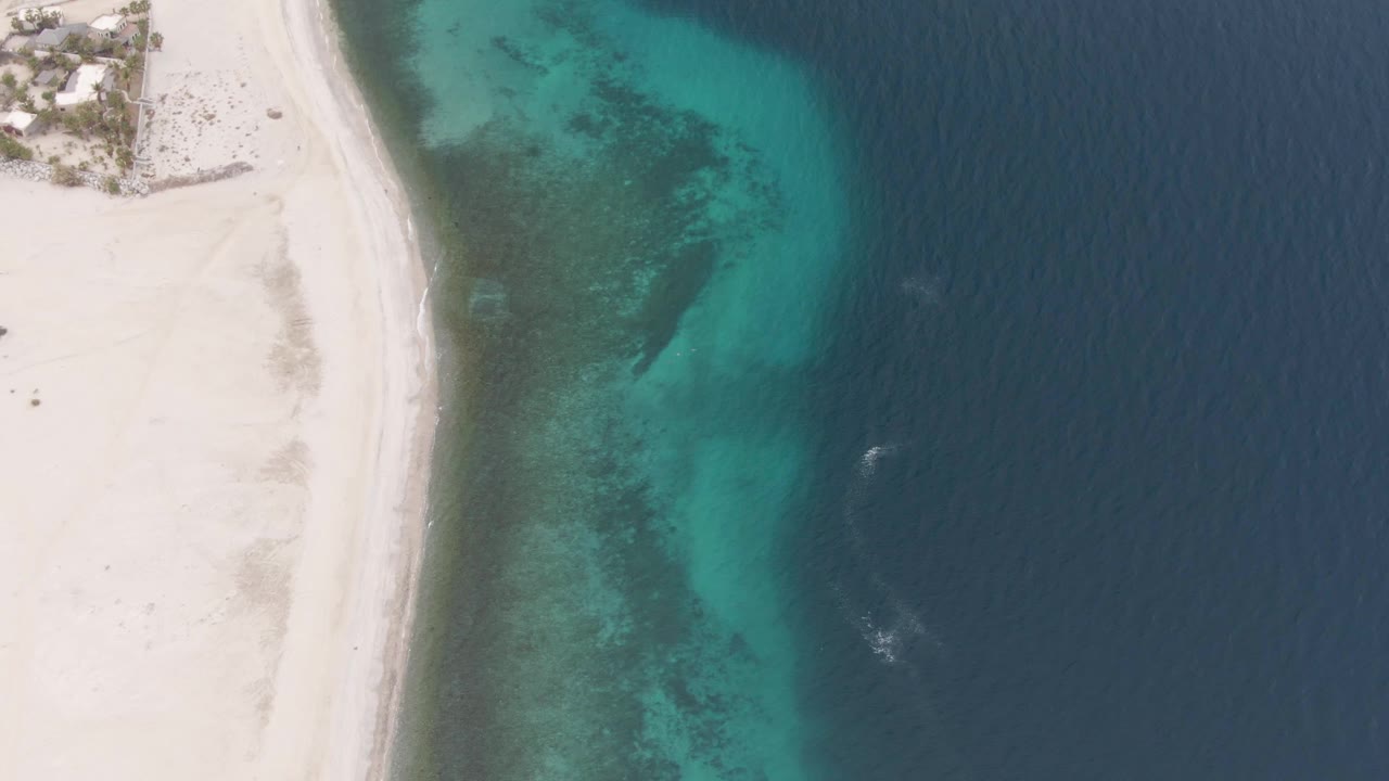 Top-down Los Barriles coast view with small structures near the upper-left sand, a curved turquoise shallows zone, deep blue water offshore, and no visible people.