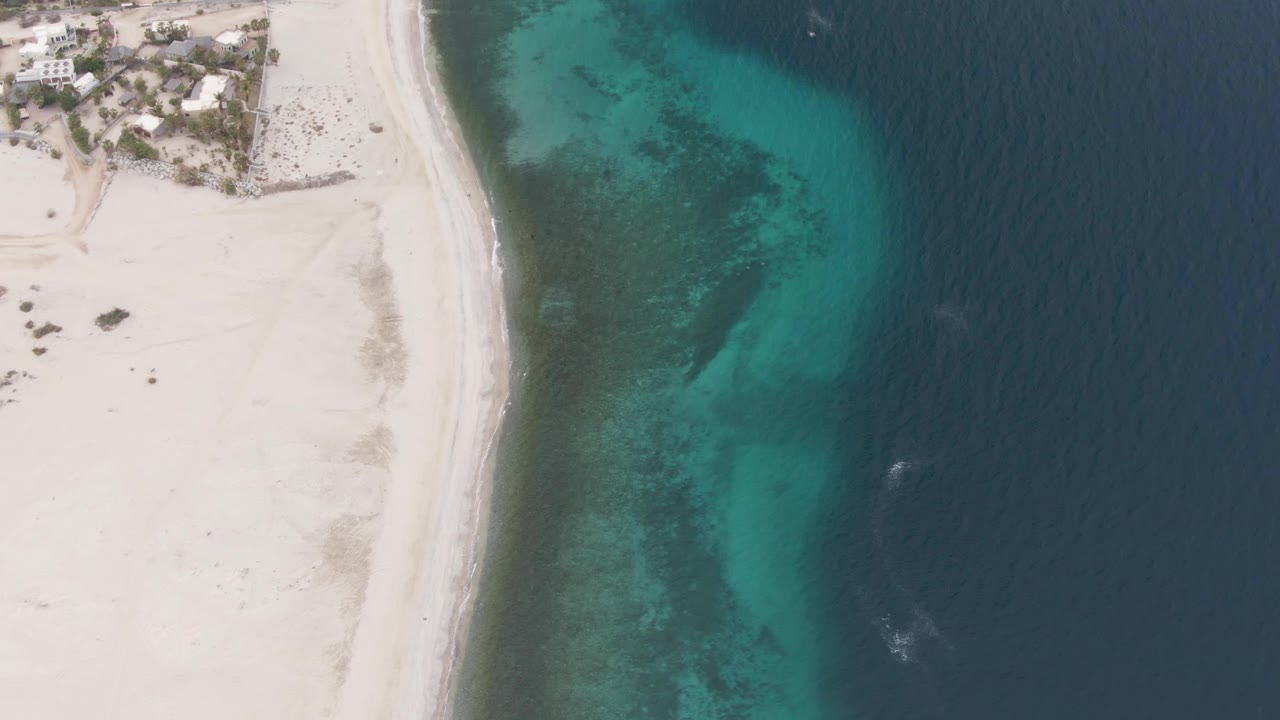 Overhead aerial view of Los Barriles beach with pale sand, coastal buildings at the upper left, turquoise shallows, darker seabed patches, deep blue water to the right, and no visible people.