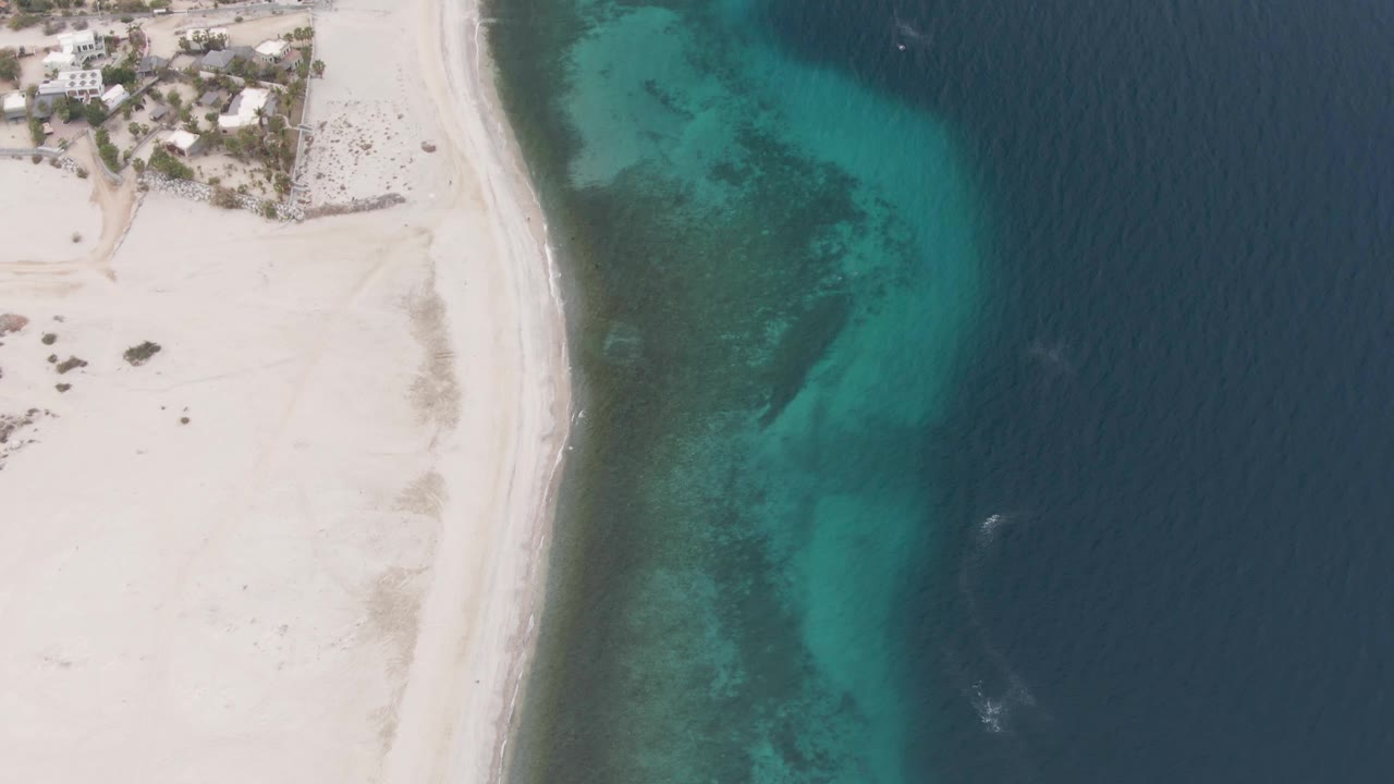 Aerial frame over Los Barriles showing the beach edge slightly farther along the coast, with turquoise water hugging the sand, deep blue ocean on the right, coastal buildings near the top, and no visible people.