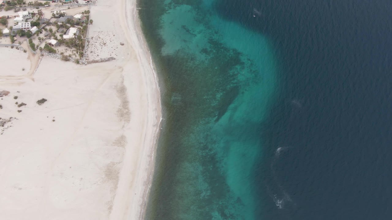 Overhead view of the Los Barriles shoreline with pale beach at left, a narrow surf line, green-blue shallow water over visible seabed, darker open ocean at right, and no visible people.