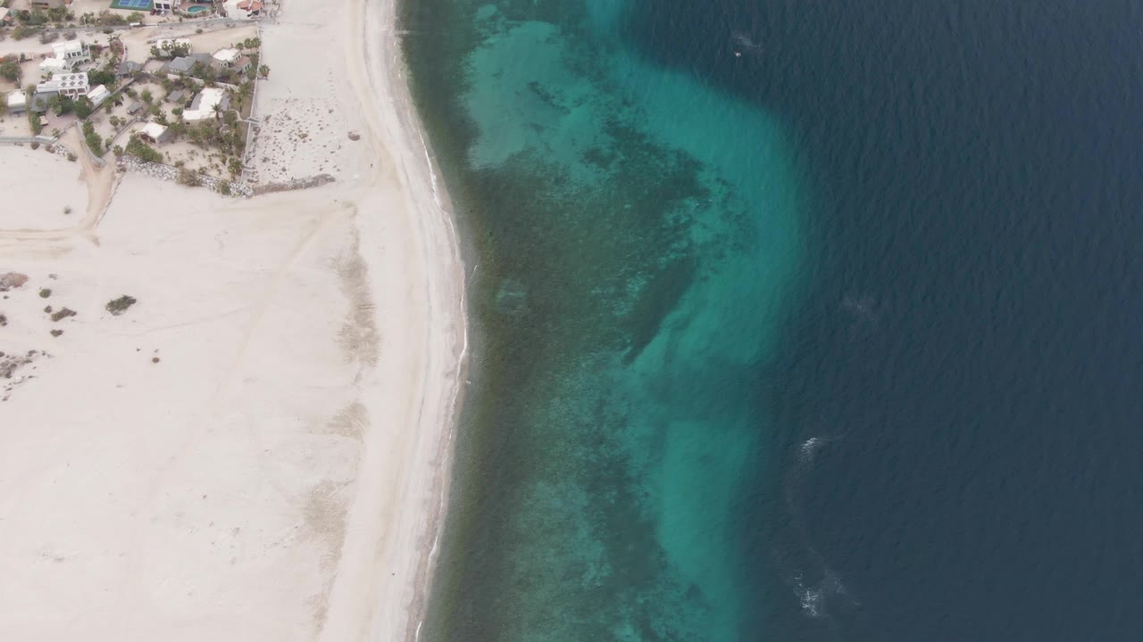 Aerial shoreline with beach, coastal homes, turquoise shallows, and blue ocean.