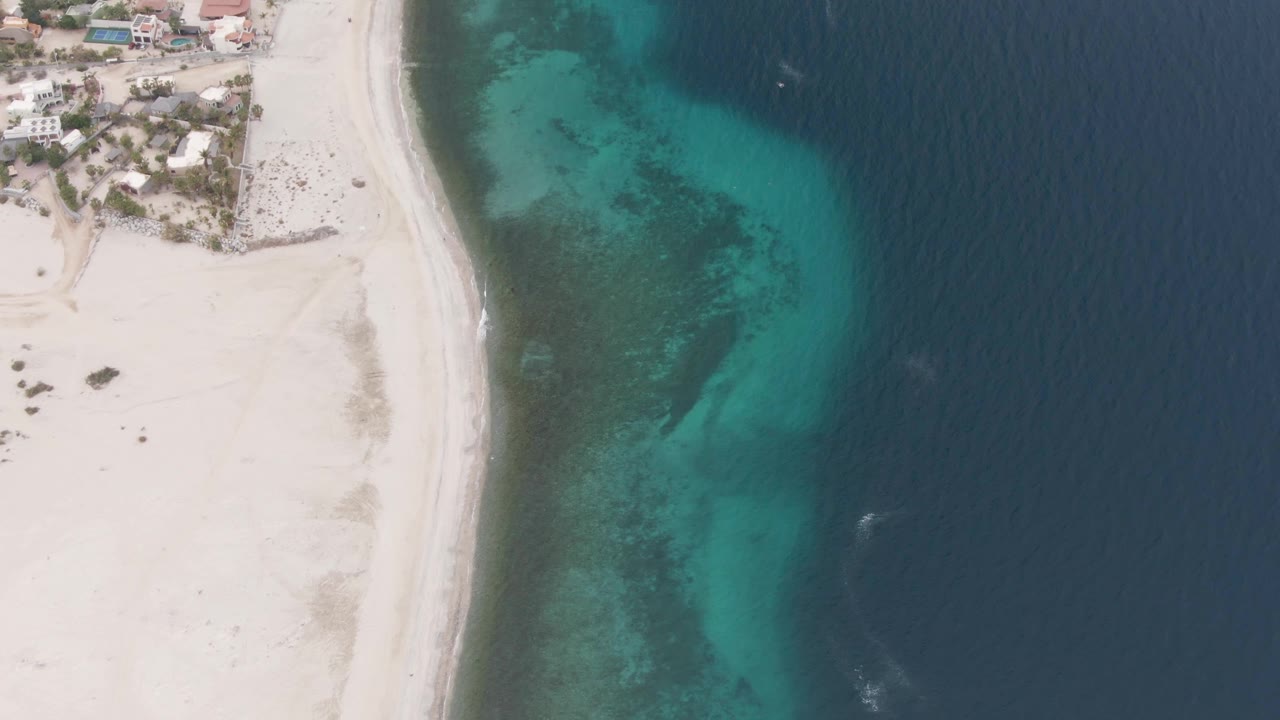 Overhead aerial frame of Los Barriles shoreline with pale sand on the left, clear turquoise shallows in the center, deep blue ocean on the right, beachfront buildings near the top edge, and no visible people.