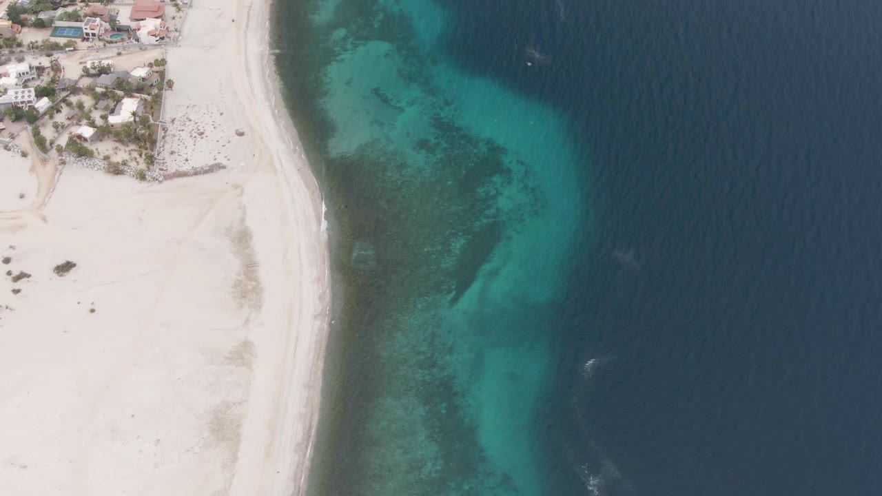 Aerial view over Los Barriles with the curved sandy beach along the left side, blue-green shallows showing darker seabed patches, deeper ocean filling the right side, and no visible people.