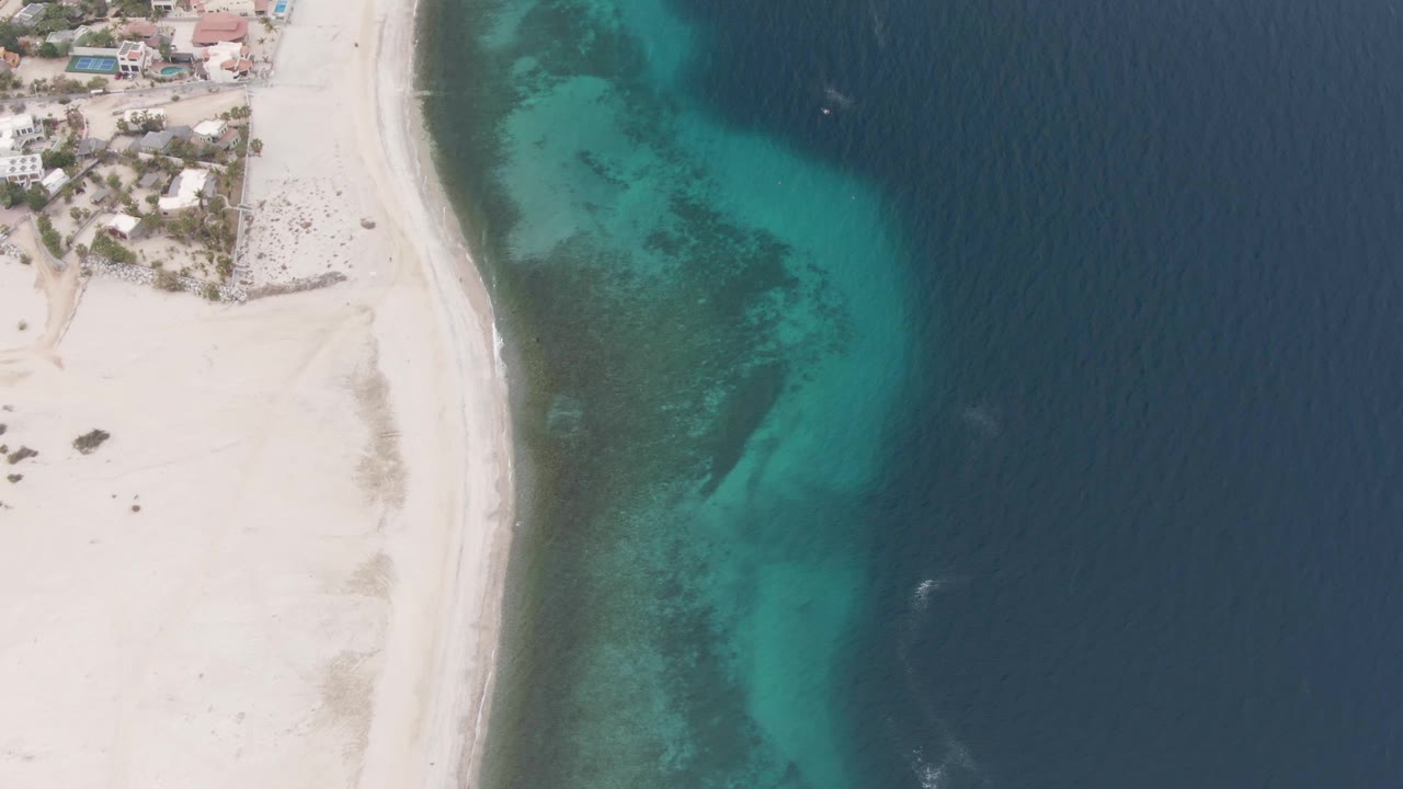 High aerial frame of the Los Barriles coast showing pale sand, a narrow surf line, turquoise water over darker underwater patches, darker open sea to the right, nearby buildings, and no visible people.