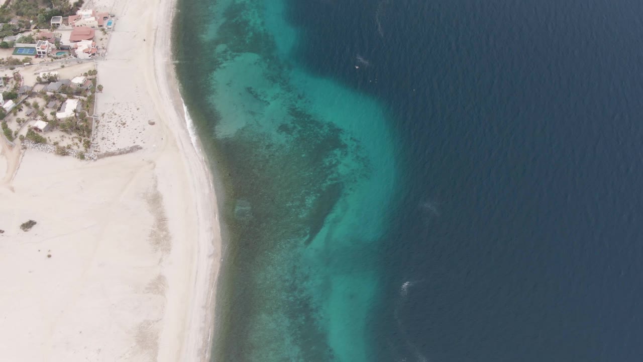 Aerial frame over Los Barriles, Mexico showing pale sand on the left, coastal buildings near the top, turquoise shallows in the center, deep blue water on the right, and no visible people.