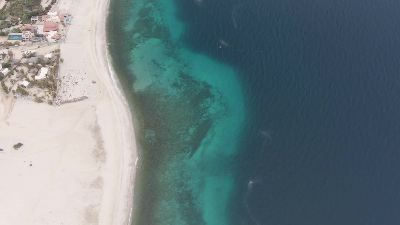Los Barriles aerial frame showing more open deep-blue ocean to the right, turquoise water along the shore, coastal houses at the upper left, pale beach below them, and no visible people.