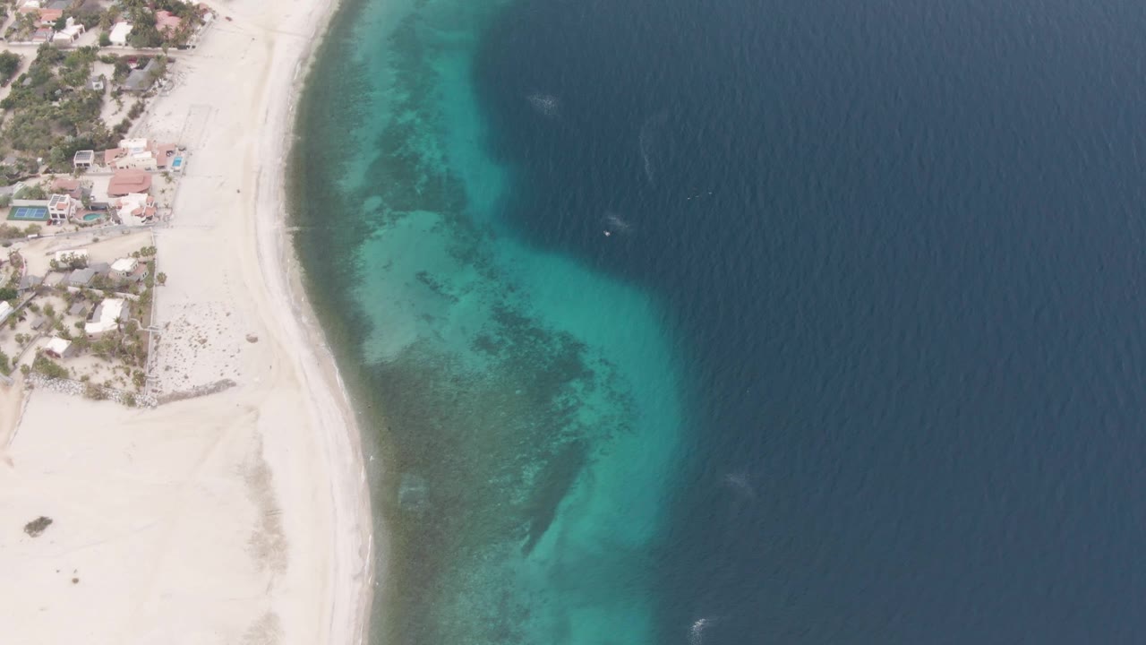 Overhead view in Los Barriles, Baja California Sur, with pale sand at left, turquoise shallows curving through the center, dark blue ocean at right, shoreline buildings near the top left, and no visible people.