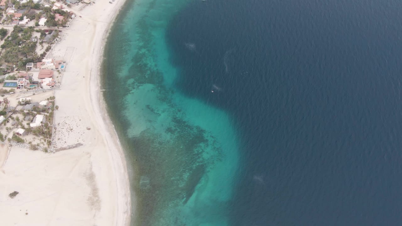 Drone view of a curved beach beside clear turquoise water and darker ocean.