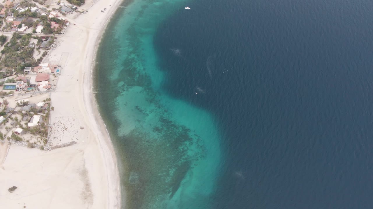 Los Barriles aerial still with white sand and houses at the upper left, transparent green-blue shallows along the beach curve, dark open water on the right, a small white boat near the top right, and no visible people.