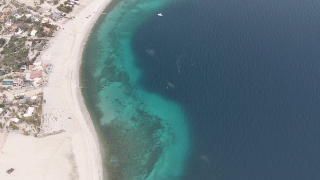 Aerial beach curve with shoreline homes, pale sand, turquoise water, and small boats offshore.