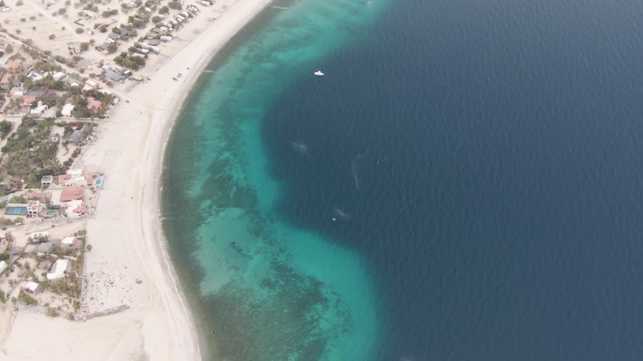 Overhead view of Los Barriles shoreline with a curved white sand beach, turquoise shallow water, darker open sea, a small white boat near the upper water, and no visible people.