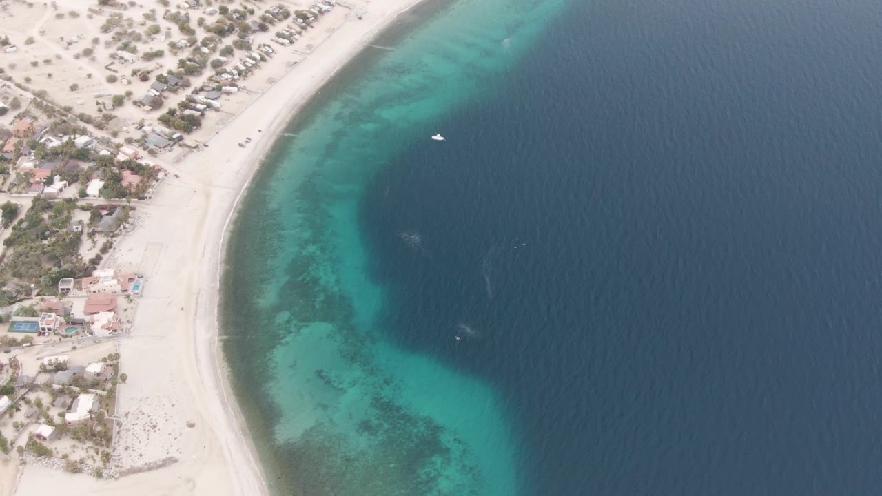 Overhead Los Barriles beach frame with the shoreline running diagonally, pale sand and buildings at left, clear turquoise shallows near shore, a small white boat in darker water, and no visible people.